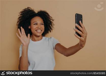 Hello  Beautiful cheerful dark skinned woman smiling at camera on modern smartphone, taking selfie on mobile phone and feeling happy, gesturing hi while making video call, isolated over studio wall. Cheerful african woman gesturing hi while making video call or taking selfie on smartphone