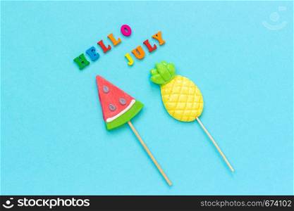 Hello July colorful text, pineapple and watermelon lollipops on stick on blue paper background. Concept vacation or holidays Creative Top view Template Greeting card, postcard.. Hello July colorful text, pineapple and watermelon lollipops on stick on blue paper background. Concept vacation or holidays Creative Top view Template Greeting card, postcard
