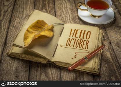 Hello October handwriting in a retro journal with decked edge handmade paper pages and a stylish pen on a rustic wooden table with a cup of tea, fall season and journaling concept