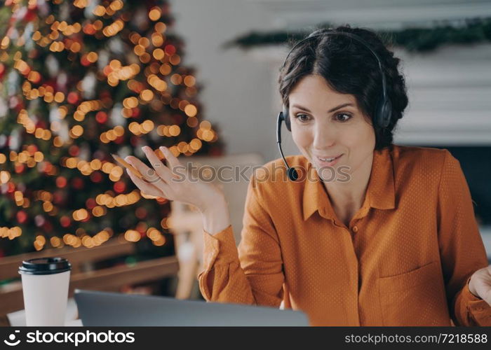 Helpful call center consultant beautiful hispanic woman in headset with mic communicates with customer while working remotely online during Christmas holidays, sitting at desk against xmas tree. Callcenter female consultant in headset talking with customer while working during Christmas holidays