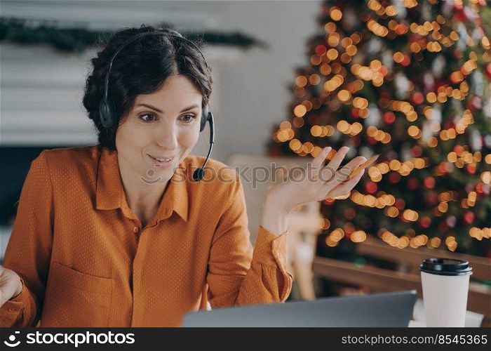 Helpful call center consultant beautiful hispanic woman in headset with mic communicates with customer while working remotely online during Christmas holidays, sitting at desk against xmas tree. Callcenter female consultant in headset talking with customer while working during Christmas holidays