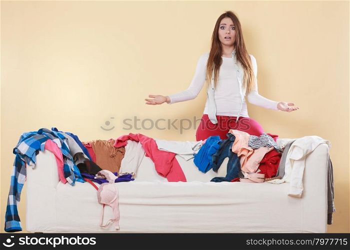Helpless woman sitting on sofa in messy room home.. Helpless woman standing behind on sofa couch in messy living room shrugging. Young girl surrounded by many stack of clothes. Disorder and mess at home.