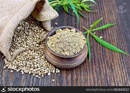 Hemp flour in a clay bowl, the grain in the bag and on the table, green cannabis leaf on a background of wooden boards