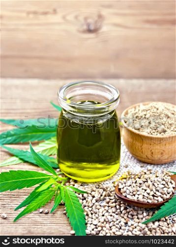 Hemp oil in a glass jar, grain in a spoon and flour in a bowl on sackcloth, cannabis leaves on a wooden board background
