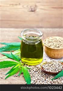 Hemp oil in a glass jar, grain in a spoon and flour in a bowl on sackcloth, cannabis leaves on a wooden board background