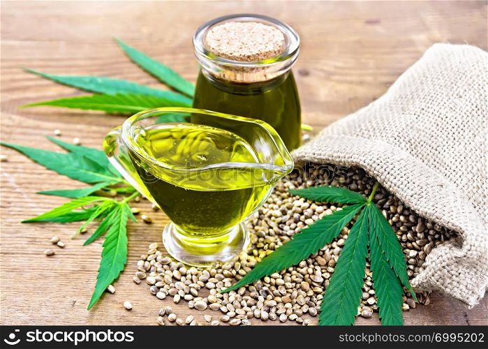 Hemp oil in a glass sauceboat and a jar with grain in a bag, leaves and stalks of cannabis on a wooden board background