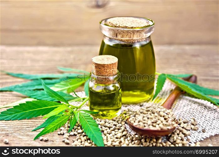Hemp oil in two glass jars, a spoon with grains on a napkin of burlap, leaves and stalks of cannabis against the background of an old wooden board