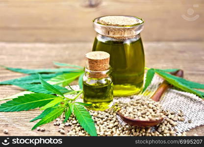 Hemp oil in two glass jars, a spoon with grains on a napkin of burlap, leaves and stalks of cannabis against the background of an old wooden board