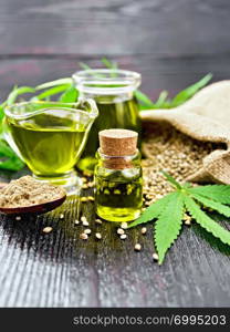 Hemp oil in two glass jars and sauceboat with grain in a sack, a spoon with flour, leaves and stalks of cannabis on a dark wooden board background