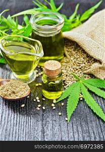 Hemp oil in two glass jars and sauceboat with grain in the bag, leaves and stalks of cannabis, a spoon with flour on the background of black wooden boards