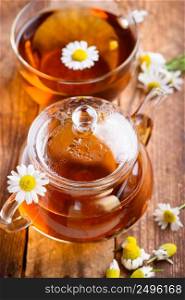 Herbal chamomile tea in cup and glass teapot with fresh chamomile herb flowers on vintage wooden table background
