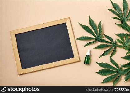 herbal medicine and aroma therapy concept - close up of hemp essential oil in glass bottle, chalkboard and cannabis leaves on beige background. hemp essential oil, chalkboard and cannabis leaves