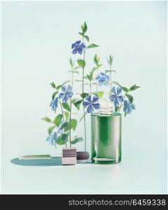 Herbal skin care cosmetics and beauty concept. Green facial Serum or oil bottle with dropper or pipette and medical flowers and herbs stand at blue background, front view.