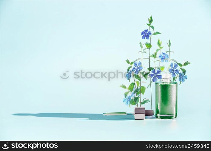 Herbal skin care cosmetics and beauty concept. Green Facial Serum or oil bottle with dropper or pipette and medical flowers and herbs stand at blue background, front view. Minimal creative layout