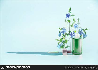 Herbal skin care cosmetics and beauty concept. Green Facial Serum or oil bottle with dropper or pipette and medical flowers and herbs stand at blue background, front view. Minimal creative layout