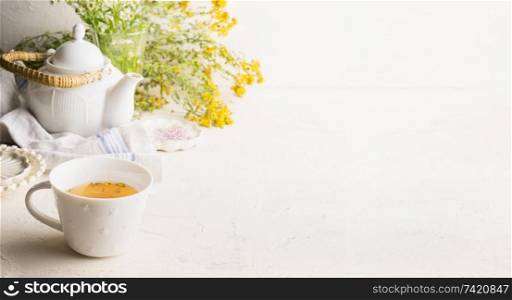 Herbal tea background with cup with yellow tea , tea pot and fresh herbs and flowers on white table at wall. Organic Tutsan tea. Wild medicinal plant concept. Copy space for your design or product.