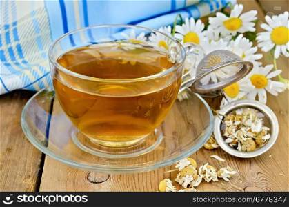 Herbal tea in a glass cup, a metal strainer with dried chamomile flowers, fresh flowers daisies, napkin against a wooden board