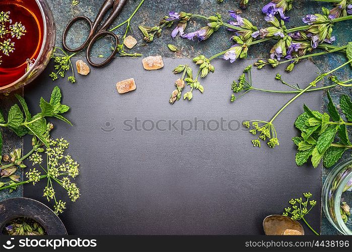 Herbal tea preparation with fresh herbs and flowers on black chalkboard background, top view, frame