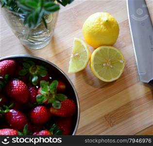 herbs in a glass, strawberries in a colander, and sliced lemons on a wood cutting board with a knife