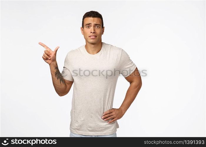 Hesitant upset or displeased young good-looking tanned man with strong muscline shape, hold hand on waist, frowning sad and indecisive, standing skeptical white background, pointing left unsure.. Hesitant upset or displeased young good-looking tanned man with strong muscline shape, hold hand on waist, frowning sad and indecisive, standing skeptical white background, pointing left unsure
