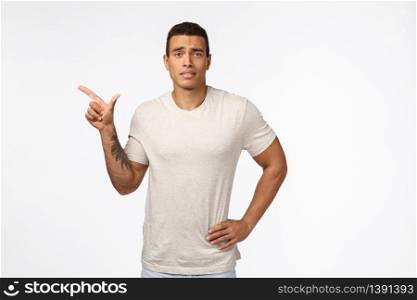 Hesitant upset or displeased young good-looking tanned man with strong muscline shape, hold hand on waist, frowning sad and indecisive, standing skeptical white background, pointing left unsure.. Hesitant upset or displeased young good-looking tanned man with strong muscline shape, hold hand on waist, frowning sad and indecisive, standing skeptical white background, pointing left unsure