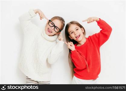 Hey, look at us, we are cool! Happy small female children in warm knitted sweaters, make grimace and have funny expressions, isolated over white background. Childhood and friendship concept.