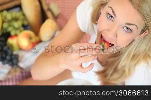 High amgle view of a beautiful blonde woman sitting on a rug at a picnic enjoying eating a strawberry