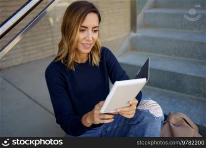 High angle of happy female in stylish jeans and sweater smiling and reading interesting e book while sitting on steps outside modern building. Positive female smiling and reading e book