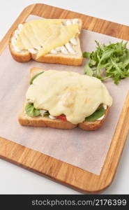 High angle of wooden board with parchment paper and toasted bread slices with cheese and vegetables and sauce before combining into delicious sandwich.. Preparation of tasty sandwich with cheese
