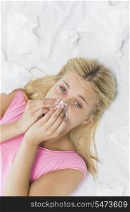 High angle portrait of young woman blowing nose in tissue paper while lying on bed