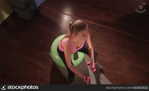High-angle shot of fit woman exercising on fitness ball with light weight dumbbells in gym