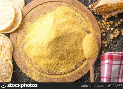 High angle view image with a pile of corn flour on a round wooden board, surrounded by corn cob, grains and puffed corn cakes, on a black wooden table.