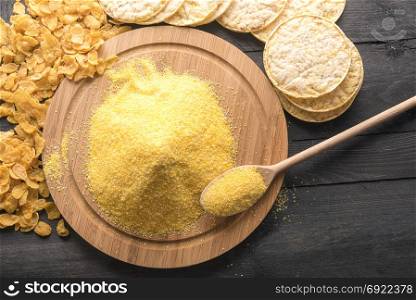 High angle view of a round wooden board full of corn flour, surrounded by products made of it, corn flakes and puffed corn cakes, on a black wooden table.