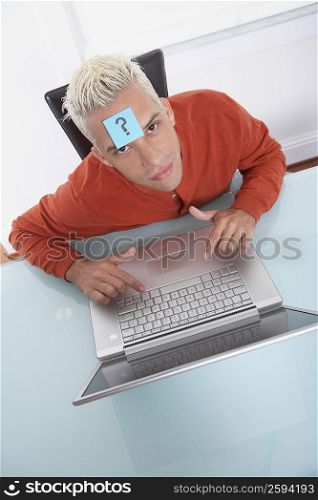 High angle view of a young man in front of a laptop with an adhesive note on his forehead