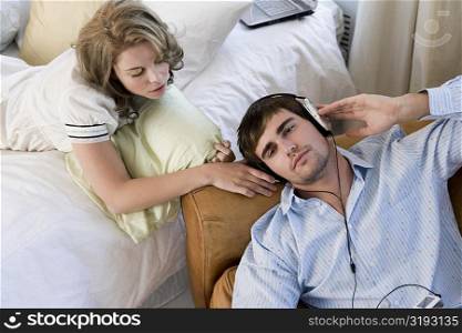 High angle view of a young man listening to an MP3 player with a young woman lying on the bed