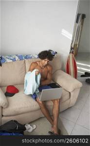 High angle view of a young man taking his t-shirt off with a laptop on his lap