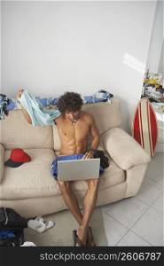 High angle view of a young man taking his t-shirt off with a laptop on his lap
