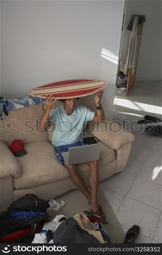 High angle view of a young man with a laptop on his lap and holding a surfboard on his head