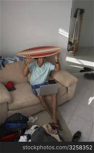 High angle view of a young man with a laptop on his lap and holding a surfboard on his head
