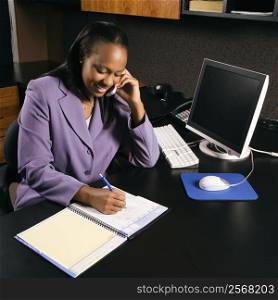 High angle view of African-American young adult business woman talking on cell phone and writing in planner in office.