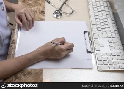 High angle view of Asian female doctor or nurse is writing something on clipboard with stethoscope and part of computer keyboard on the table at hospital office room