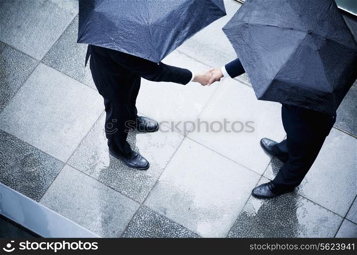 High angle view of two businessmen holding umbrellas and shaking hands in the rain