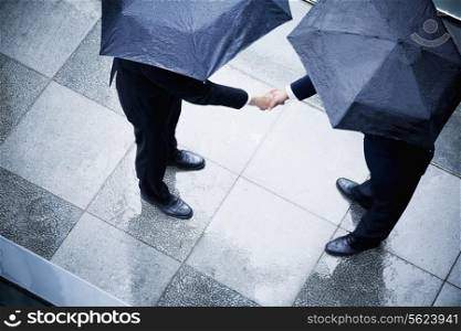 High angle view of two businessmen holding umbrellas and shaking hands in the rain