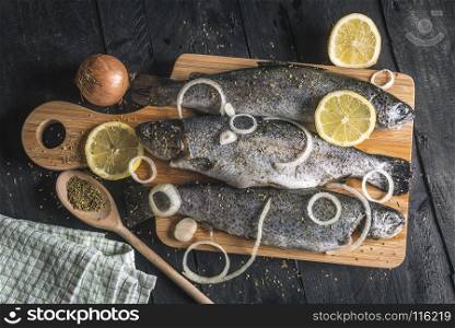 High angle view with uncooked fish on a wooden cutting board, seasoned with dried herbs, lemon slices and onion rings, on a vintage black table.