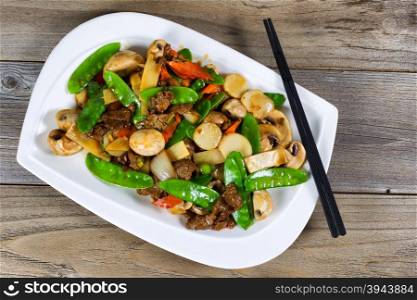 High angled view of Asian dish consisting of sliced juicy beef rice, onion, mushroom, green peas, and red pepper. Chopsticks on side of plate with rustic wood underneath.