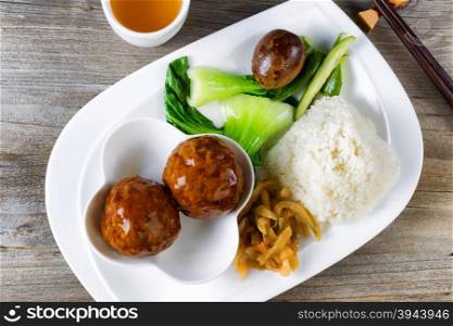 High angled view of Asian saucy meatballs, rice, egg, cucumber and bok choy on white plate. Chopsticks and green tea in background.