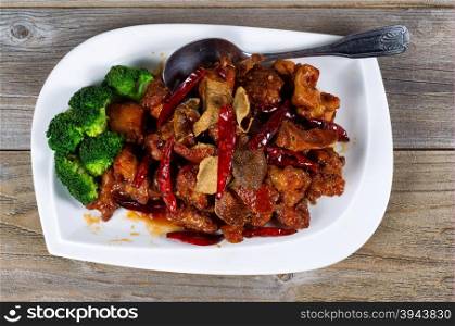 High angled view of Chinese dish consisting of fried tofu, red peppers, and broccoli. Large serving spoon on side of plate with rustic wood underneath.