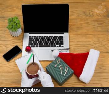 High angled view of hands wearing white gloves while preparing to drink hot chocolate with computer, present, cap, notepad, pencil and plant on desktop. Christmas concept of Santa Claus office.