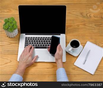 High angled view of male hand holding cell phone while typing on computer keyboard on desktop.