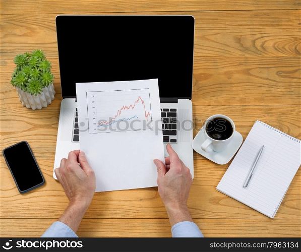 High angled view of male hands holding data graph with computer, coffee, notepad, pen and plant on desktop.&#xA;&#xA;